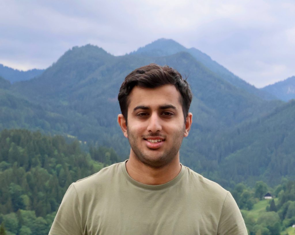 Umer smiling in front of green forested mountains beneath a cloudy sky.